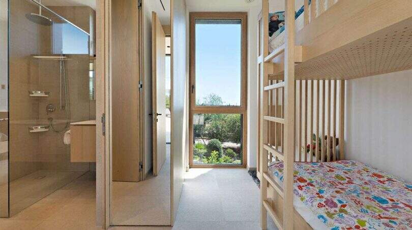 Children's room with wooden bunk bed and colorful bedding, adjacent modern glass bathroom with rain shower, garden view.