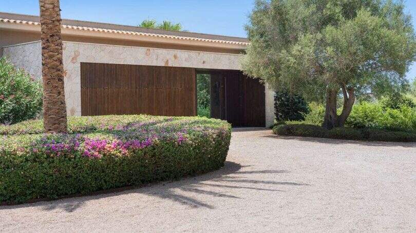 Entrance area of a modern villa in S'Alqueria Blanca with wooden gate, natural stone wall and Mediterranean garden.