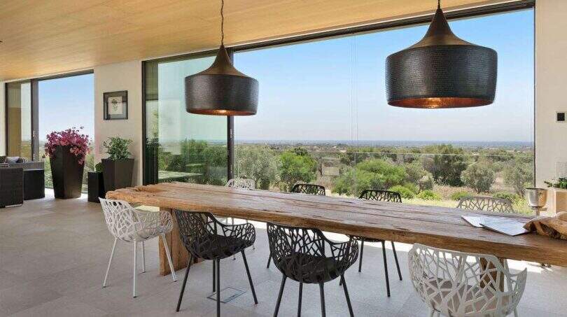 Open dining area with rustic wooden table, modern designer chairs and panoramic windows with sea view in S'Alqueria Blanca.