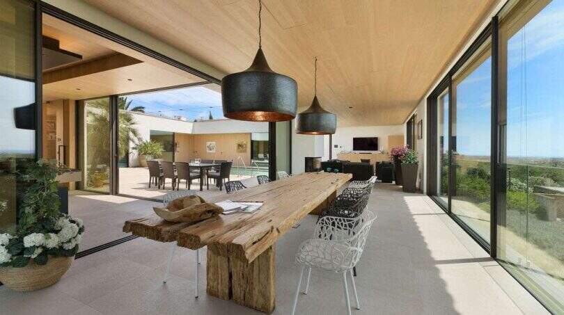 Covered terrace with rustic wooden table, designer chairs and panoramic glazing with sea view in S'Alqueria Blanca
