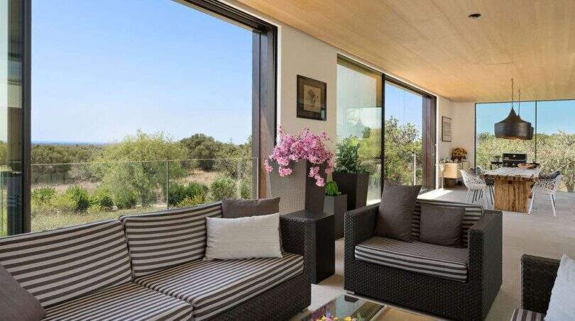 Elegant living area with panoramic windows, rattan furniture and sea view in modern design house in S'Alqueria Blanca.