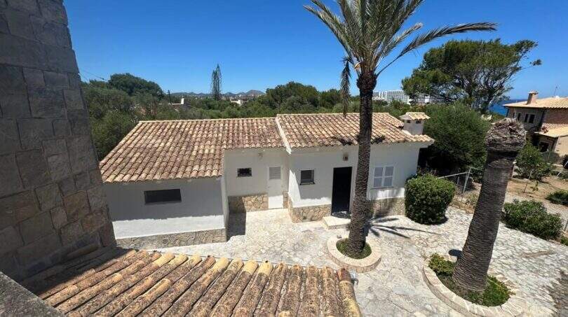 Außenansicht mediterrane Villa Cala Murada mit Terrakotta-Dach, Palme und Steinpflaster bei blauem Himmel.