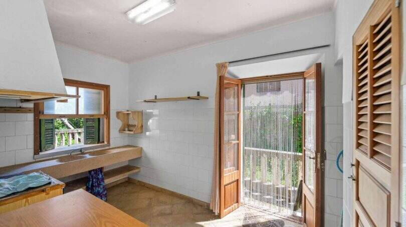 Helle Küche der Villa Cala Figuera mit Steinarbeitsplatte, Holztür zum Balkon und weißen Wandfliesen.