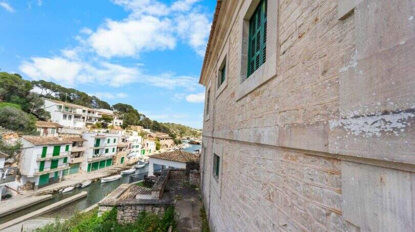 Außenansicht Villa Cala Figuera: Sandsteinmauer mit grünen Fensterläden, Blick auf malerischen Hafen mit Booten.