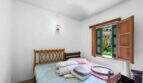 Helles Schlafzimmer der Villa Cala Figuera mit Doppelbett, Holzkopfteil und Fensterläden mit Gartenblick.