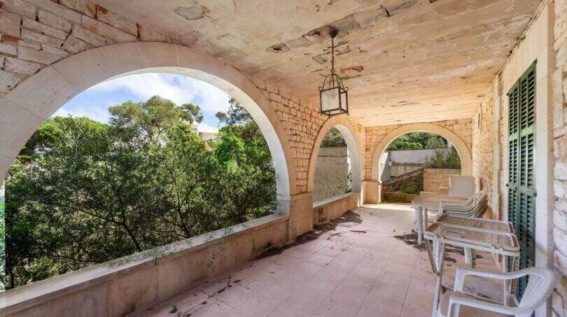 Verfallene Loggia mit Rundbögen, Natursteinwänden und weißen Gartenstühlen, Blick auf grüne Pinien in Cala Figuera