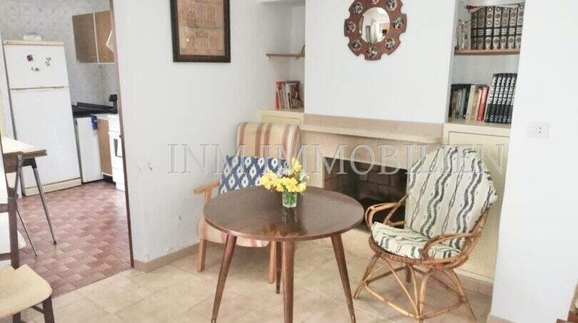 Wohnzimmer mit rundem Holztisch, Rattanstühlen, Kamin und Bücherregal, Blick in die Küche