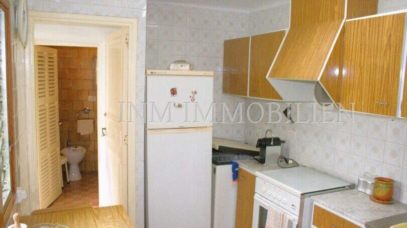 Schlichte Küche mit Holzschränken, Kühlschrank und Herd, Blick in gefliestes Bad in Dorfhaus Felanitx