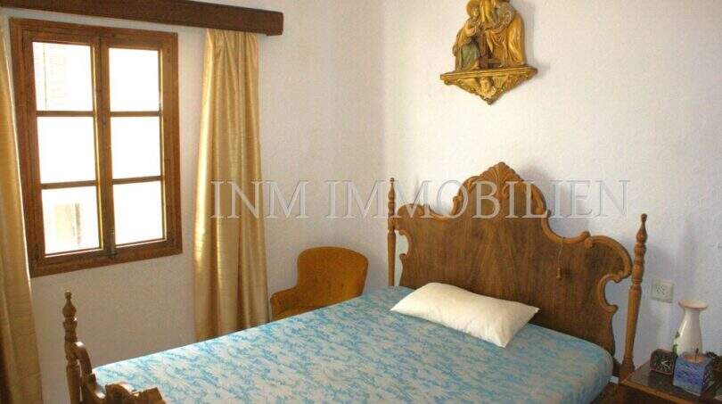 Schlafzimmer mit antikem Holzbett, religiöser Wanddekoration und kleinem Fenster mit goldenen Vorhängen in Felanitx