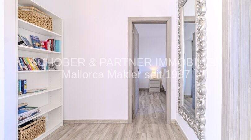 Heller Flurbereich der mediterranen Villa in Sa Coma mit weißem Bücherregal, Holzboden und elegantem Spiegel.