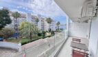 Balkon mit Glasgeländer, Gartenstuhl und Blick auf Palmen und weiße Apartmenthäuser in Palmanova