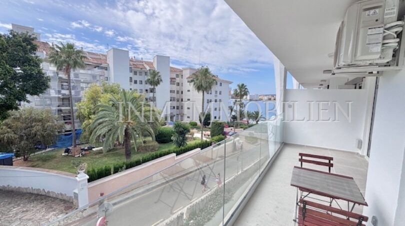 Balkon mit Glasgeländer, Gartenstuhl und Blick auf Palmen und weiße Apartmenthäuser in Palmanova