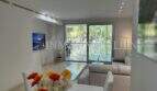 Helles Wohnzimmer der Luxuswohnung Bendinat mit Glasfront, Sofa, TV und Blick in grüne Gartenanlage.