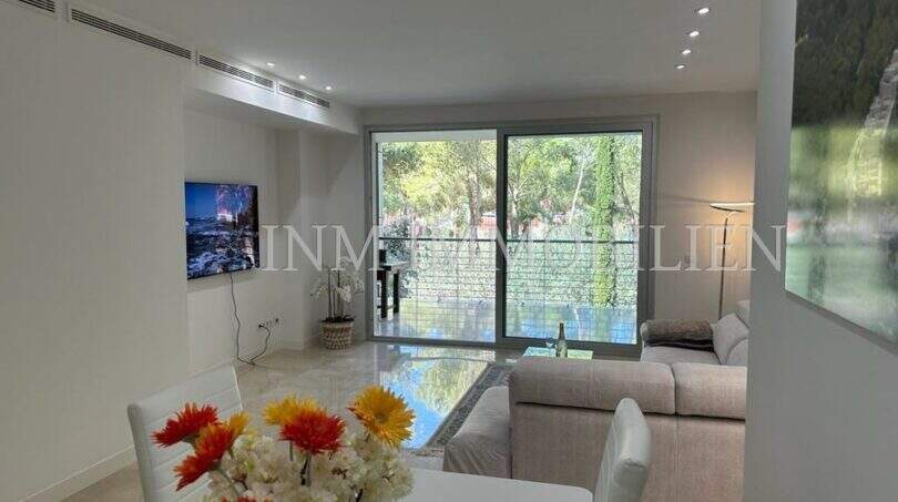 Helles Wohnzimmer der Luxuswohnung Bendinat mit Glasfront, Sofa, TV und Blick in grüne Gartenanlage.