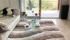 Helles Wohnzimmer mit weißem Ledersofa, Glastisch, Designerteppich und Zugang zur Terrasse mit Grünblick in Bendinat