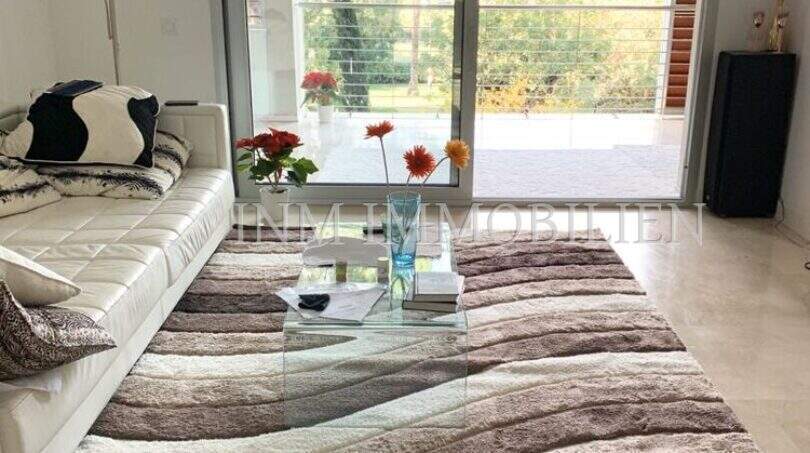 Helles Wohnzimmer mit weißem Ledersofa, Glastisch, Designerteppich und Zugang zur Terrasse mit Grünblick in Bendinat