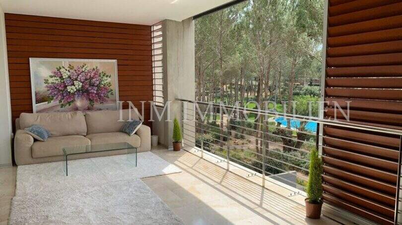 Helles Wohnzimmer mit Sofa, Holzwand und großer Glasfront mit Blick auf Pinien und Gemeinschaftspool in Bendinat