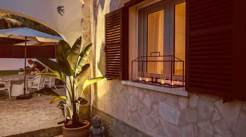 Beleuchtete Terrasse mit Buddha-Statue, Bananenpalme und Kerzen am Fenster bei Dämmerung in Cala Murada