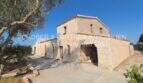 Außenansicht eines rustikalen Landhauses in Portocristo mit Natursteinwänden, Carport und mediterranem Bewuchs unter blauem Himmel.