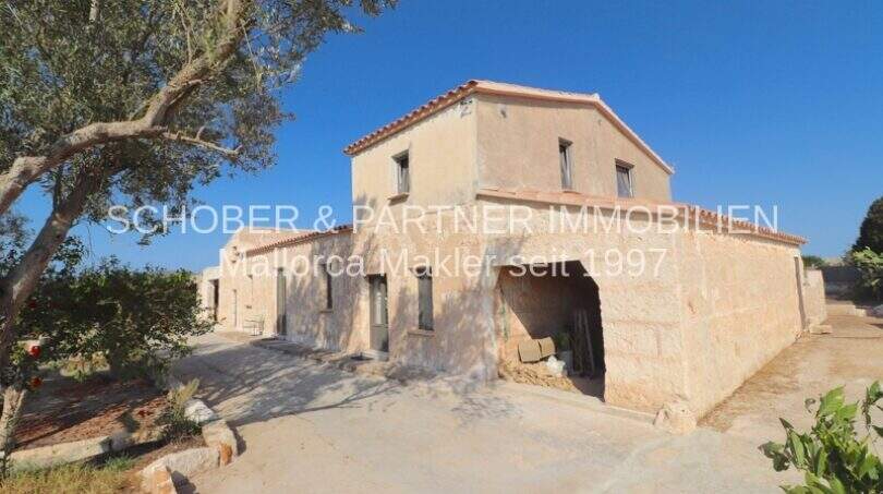 Außenansicht eines rustikalen Landhauses in Portocristo mit Natursteinwänden, Carport und mediterranem Bewuchs unter blauem Himmel.