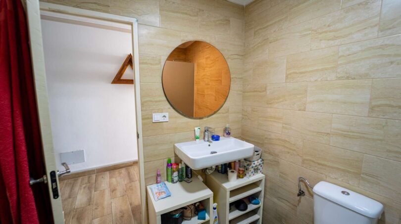Baño con espejo redondo, lavabo y azulejos de piedra claros en casa unifamiliar alquilada en Felanitx