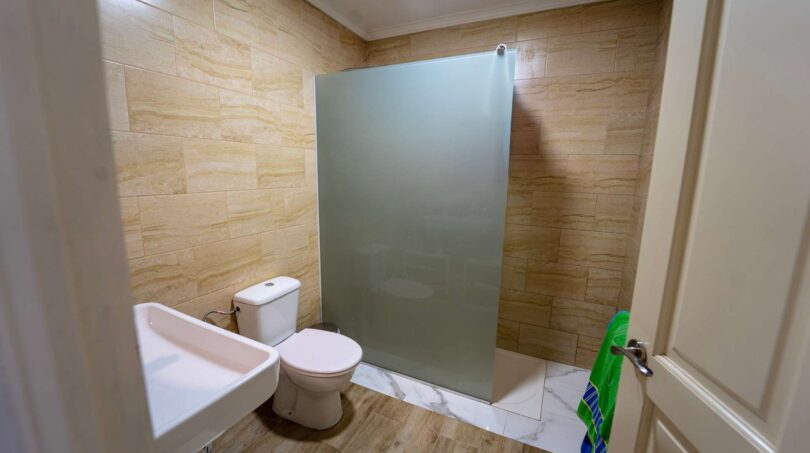 Baño con inodoro, lavabo y ducha con división de vidrio esmerilado, azulejos claros con aspecto de arenisca, casa multifamiliar Felanitx