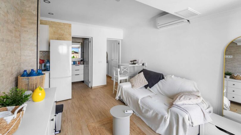 Luminosa sala de estar con sofá blanco, suelo de madera y cocina abierta en casa de múltiples familias Felanitx