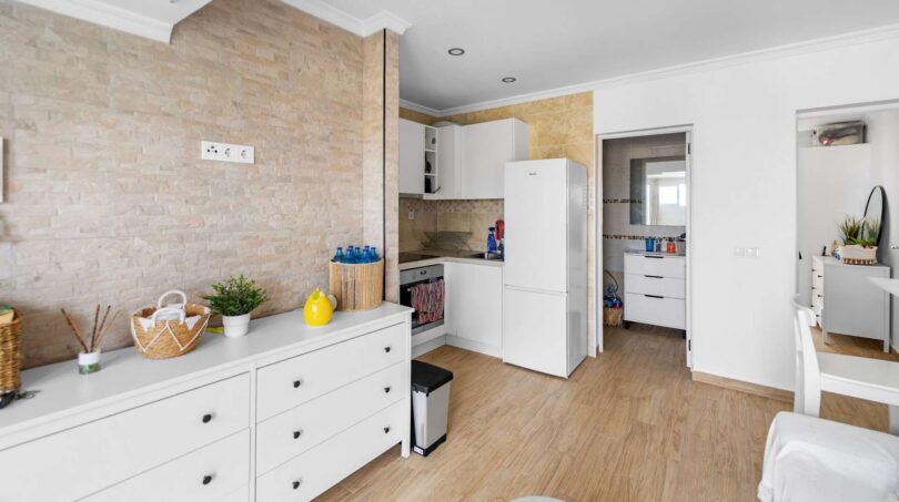 Cocina-comedor abierta con armarios blancos, revestimiento de pared de piedra y suelo de madera en casa unifamiliar alquilada en Felanitx.