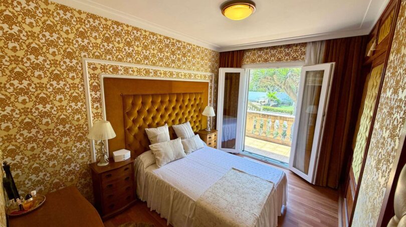 Bedroom of the retro villa in Porto Cristo with upholstered headboard, gold wallpaper and balcony with garden view.