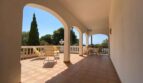 Covered terrace of the villa in Porto Cristo with round arches, lounge chairs and sea view in bright sunshine.
