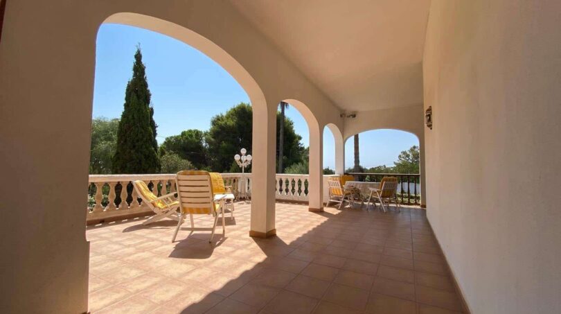 Covered terrace of the villa in Porto Cristo with round arches, lounge chairs and sea view in bright sunshine.
