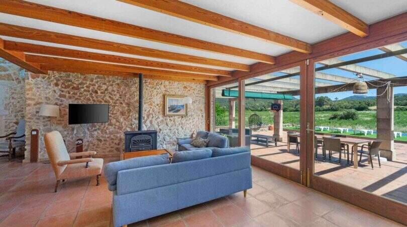 Wohnzimmer der Finca in Felanitx mit Holzbalkendecke, Steinwand, blauem Sofa und Panoramablick auf grüne Landschaft.