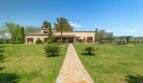 Exterior view of the Felanitx finca: well-maintained lawn, stone path, palm trees and Mediterranean farmhouse under blue sky.