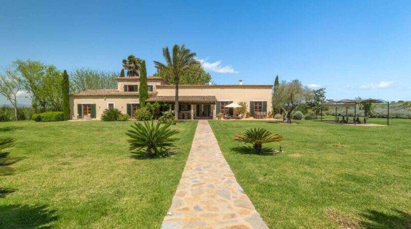Exterior view of the Felanitx finca: well-maintained lawn, stone path, palm trees and Mediterranean farmhouse under blue sky.