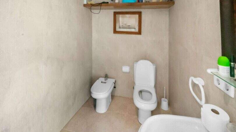 Helles Badezimmer der Wohnung in Porto Colom mit WC, Bidet, Holzregal und beigefarbenen Fliesen.