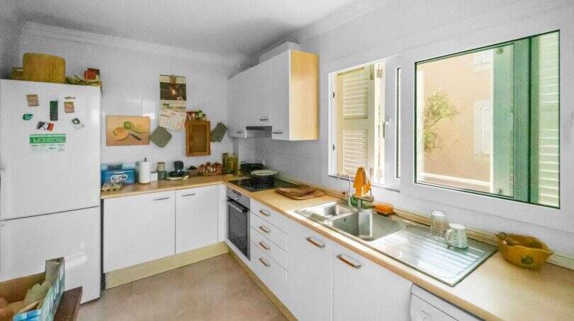 Helle Küche der Wohnung in Porto Colom mit weißen Schränken, Holzarbeitsplatten und Fenster zum Innenhof.
