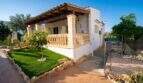 Außenansicht Villa Porto Colom: weißes Landhaus mit Holzveranda, Steinbalustrade und gepflegtem Garten mit Kunstrasenfläche.