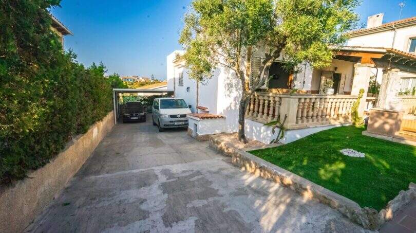 Einfahrt mit Carport und zwei Fahrzeugen, gepflegter Rasen und mediterrane Terrasse eines Einfamilienhauses in Porto Colom.