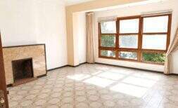 Sala de estar vacía con chimenea de mármol, piso de baldosas con patrón y grandes ventanas de madera, que requiere renovación