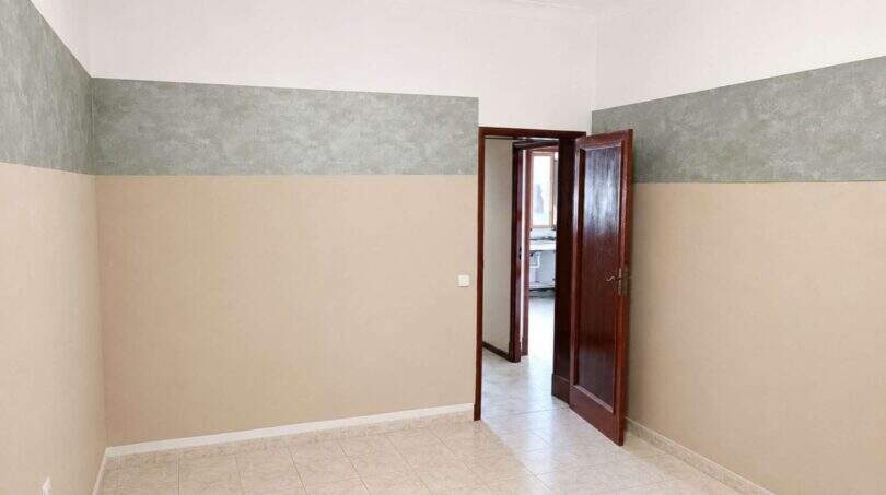 Leeres Zimmer der Altbauwohnung in Palma mit Fliesenböden, zweigetönten Wänden und Holztür zum Flur.