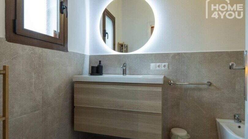 Modernes Badezimmer mit Holzwaschtisch, beleuchtetem Rundspiegel und Natursteinoptik-Fliesen im Bungalow Alcúdia