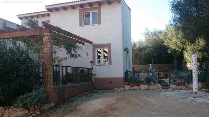Exterior view of Cala Murada chalet: white country house with wooden pergola, driveway, garden and trees in the background.