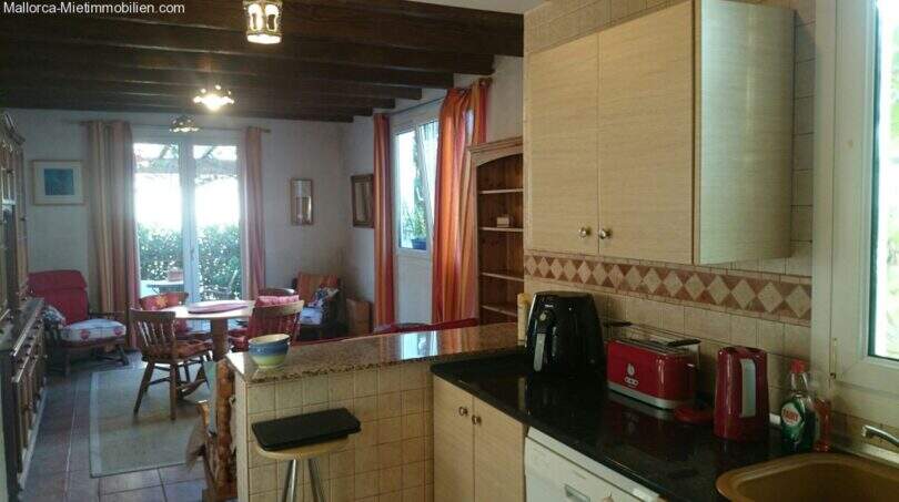 Open kitchen with dark countertop and wooden beam ceiling, adjacent living-dining area with garden view in the Cala Murada chalet