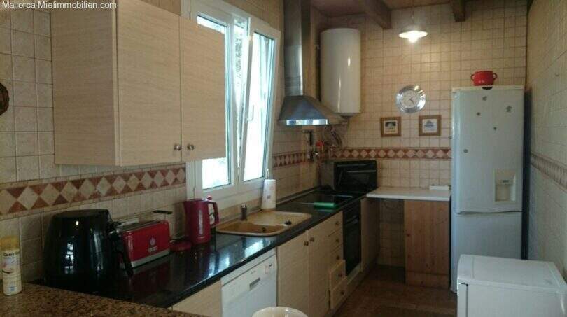 Kitchen of the chalet in Cala Murada with dark countertop, built-in appliances, tiles and forest view through the window.