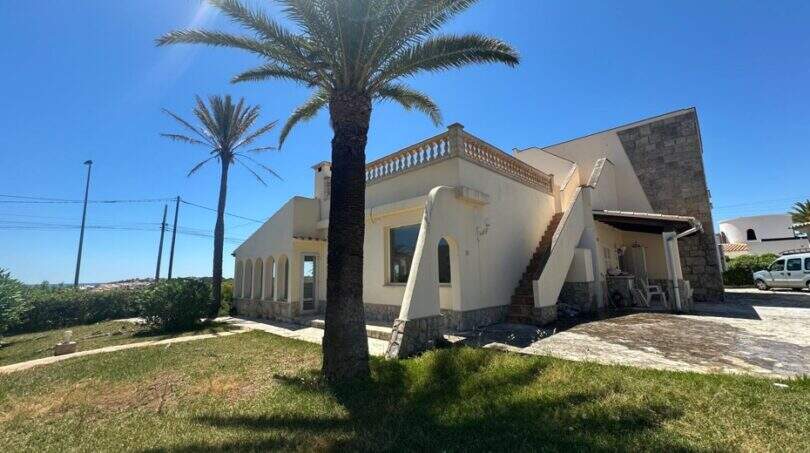 Mediterrane Villa in Cala Murada mit Palmen, Steinverkleidung, Balustrade und gepflegtem Rasen bei strahlendem Sonnenschein.