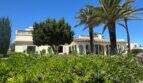 Außenansicht mediterrane Villa in Cala Murada mit Palmen, üppigem Grün und hellem Mauerwerk unter blauem Himmel.