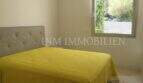 Helles Schlafzimmer mit grauem Polsterbett, gelbem Überwurf und Fenster mit Gartenblick in Luxuswohnung Bendinat