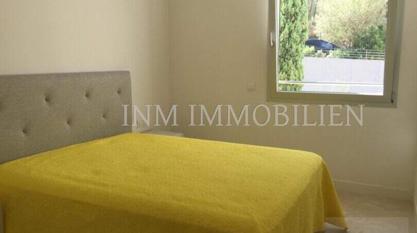 Helles Schlafzimmer mit grauem Polsterbett, gelbem Überwurf und Fenster mit Gartenblick in Luxuswohnung Bendinat