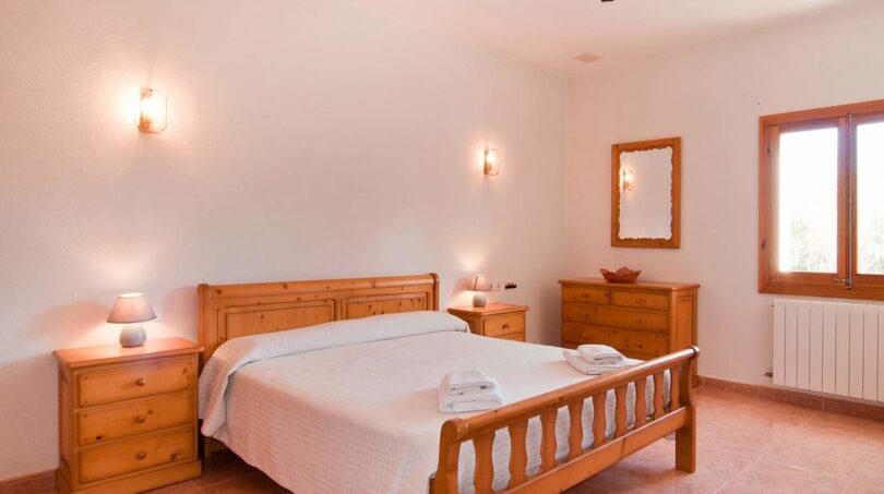 Helles Schlafzimmer der Finca mit Doppelbett, Holzmöbeln, Wandleuchten und Fenster zur Naturlandschaft.