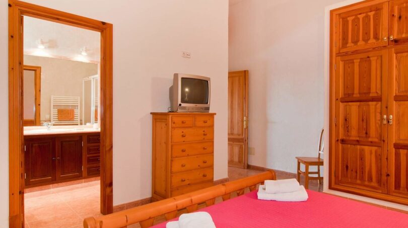 Schlafzimmer der Finca mit Holzmöbeln, CRT-TV, rosa Bett und Blick ins angrenzende Bad